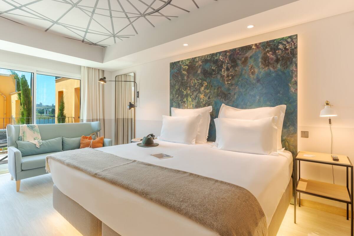 A modern hotel room with a large bed, a sofa, and a view of the outdoors.