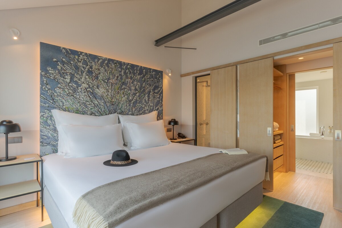 A modern hotel room with a large bed, a nightstand, a desk, a wardrobe, and a bathroom visible through an open door.