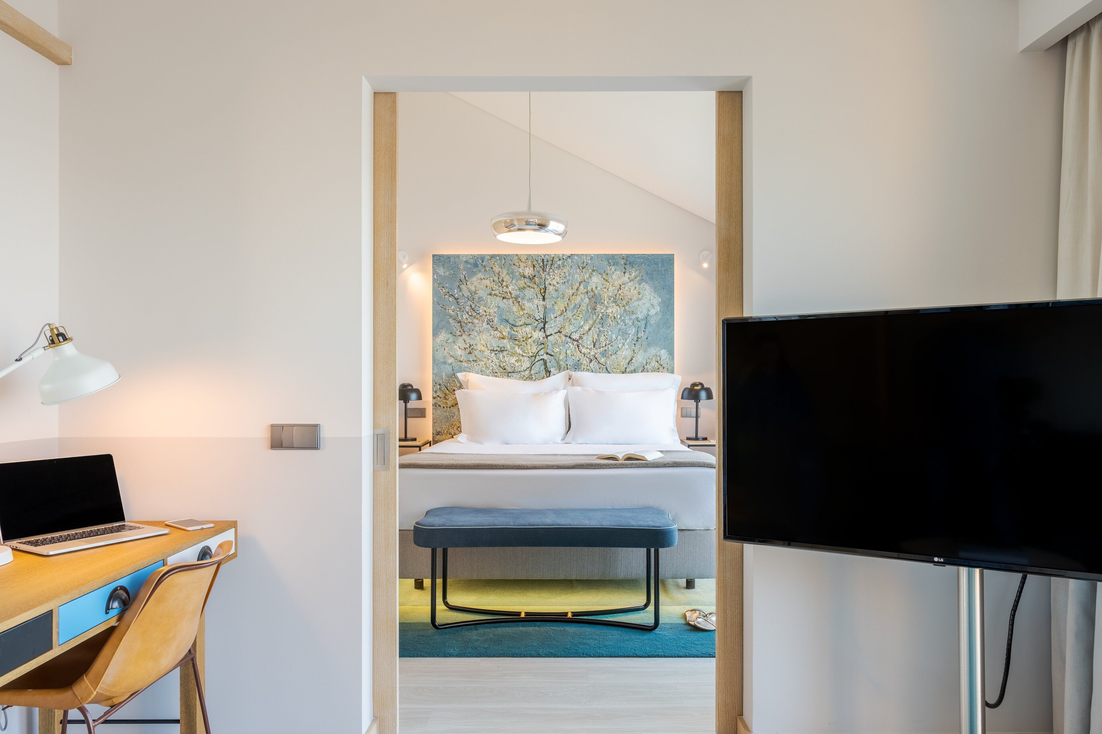 Habitación de hotel con cama, escritorio con una computadora portátil, televisor y un cuadro en la pared.