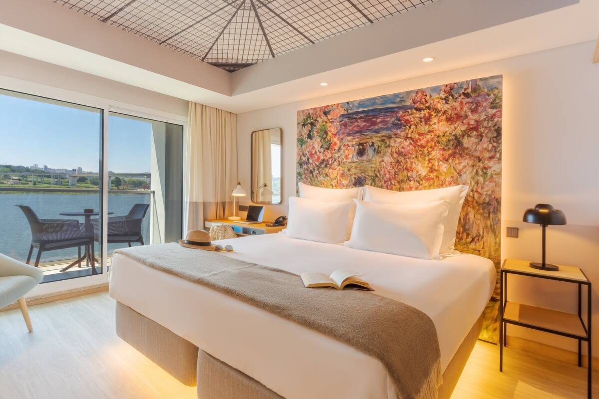 A modern hotel room with a large bed, a desk with a laptop, a chair, and a view of a body of water.