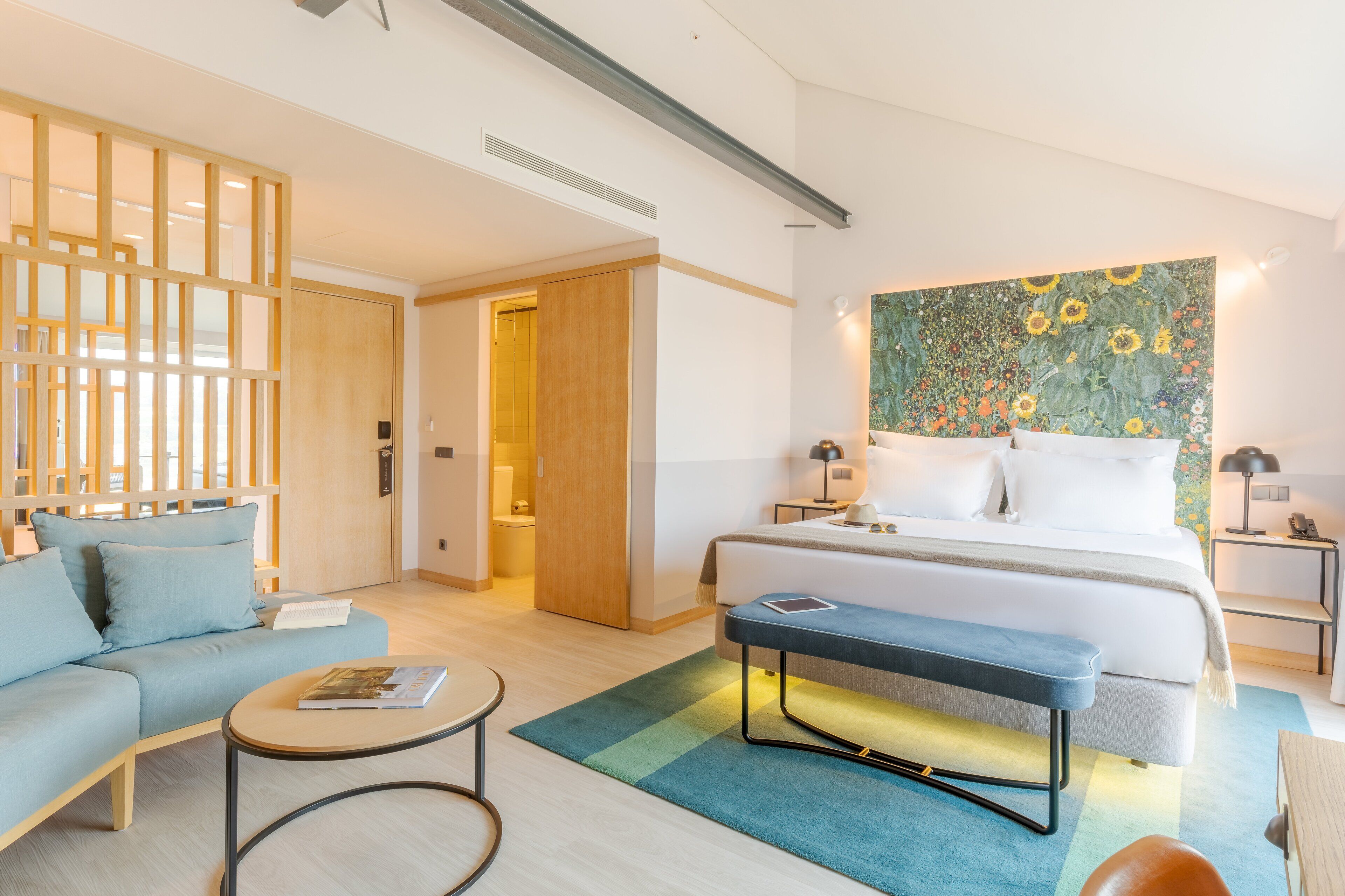 A modern hotel room with a bed, a sofa, a bench, a small table, and a wall with a floral mural.