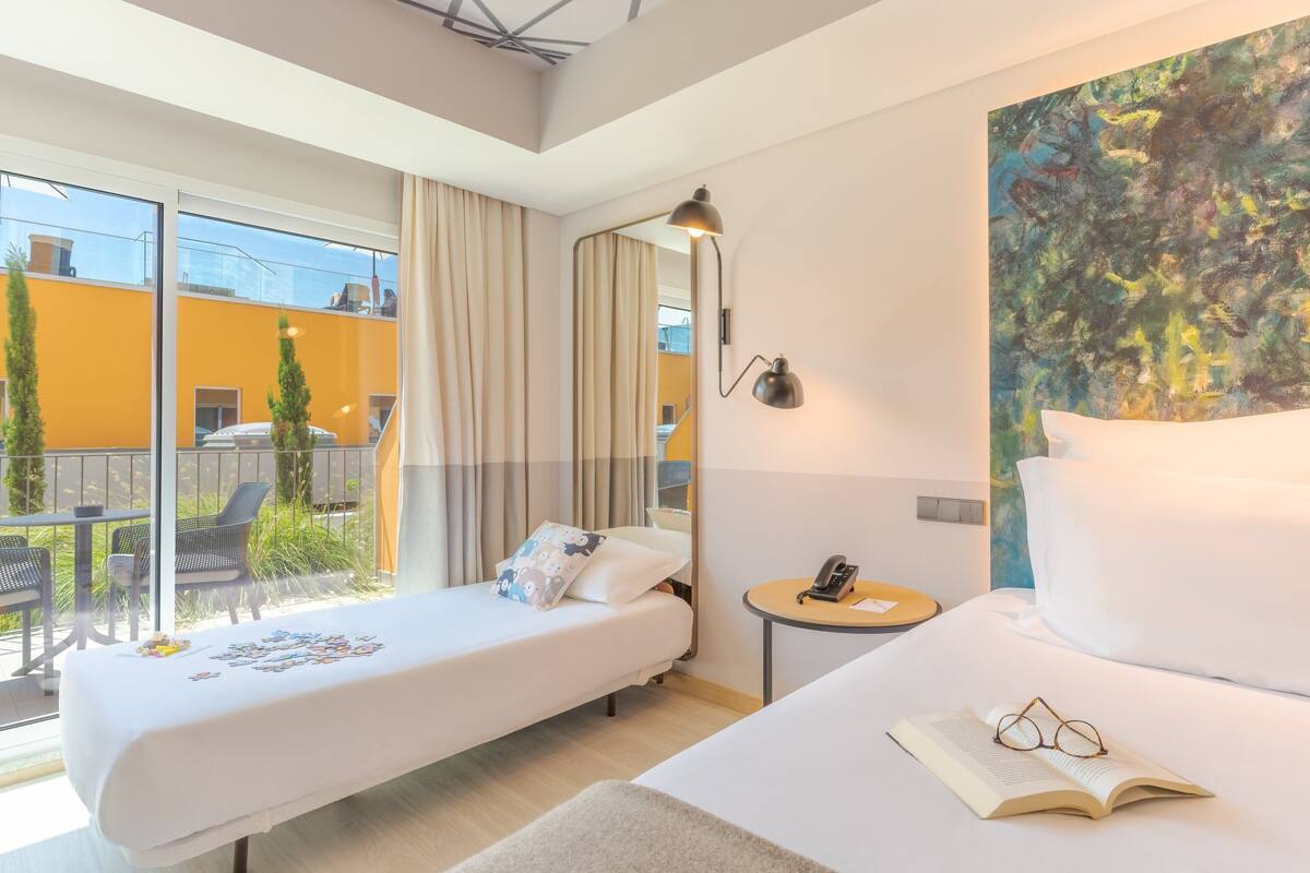 A hotel room with two beds, a balcony with outdoor seating, a painting on the wall, and a bedside table with a lamp and a phone.