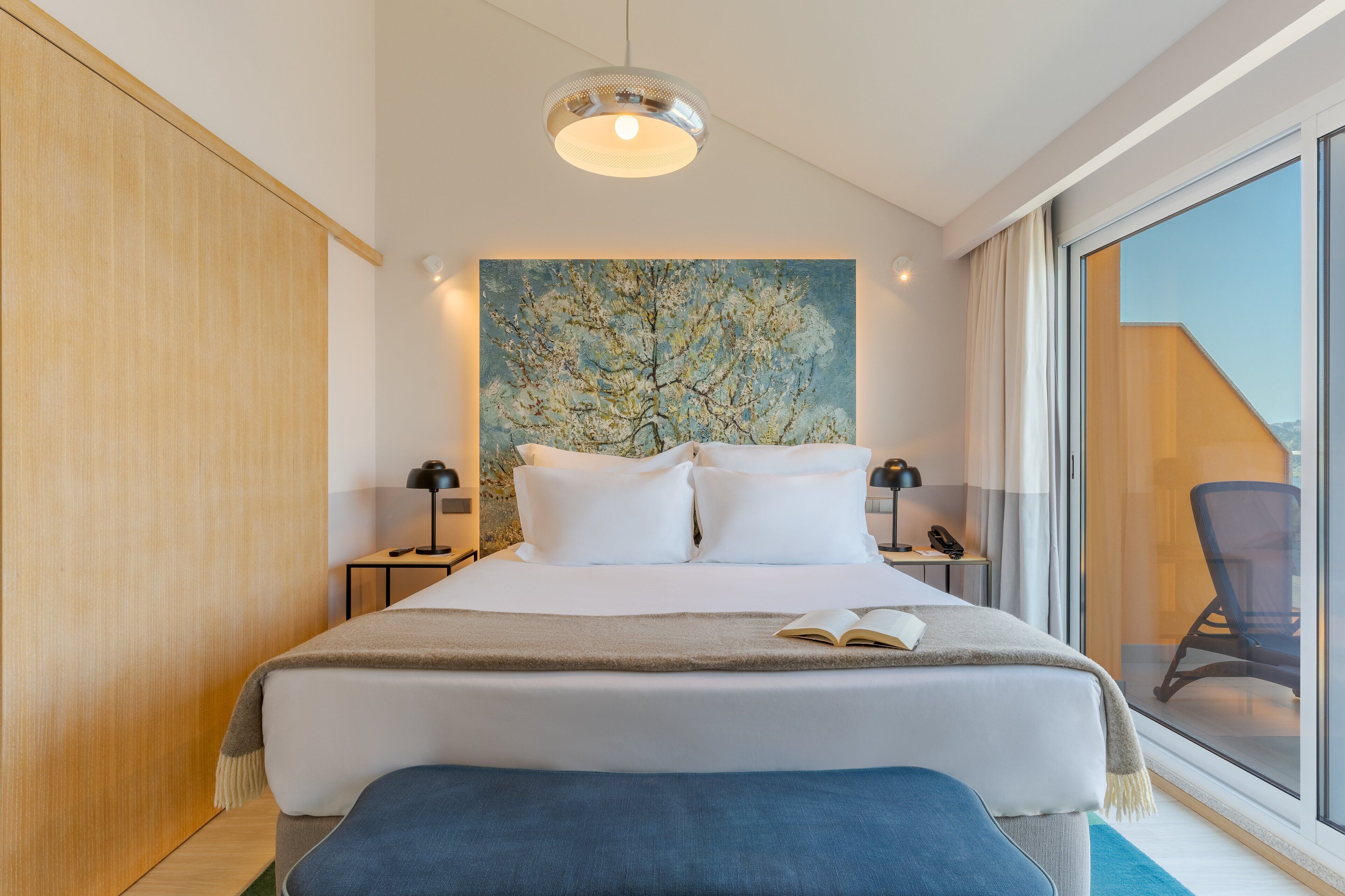 A modern hotel room with a large bed, a painting of a tree, a desk with a lamp, and a view through a glass door.