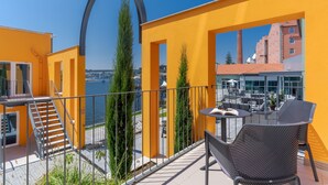 A balcony with a yellow wall, a metal railing, and a table with a chair. There are tall trees and a view of buildings in the background. - Pestana Douro Riverside - Preferred Hotels & Resorts, Lifestyle (Gondomar)