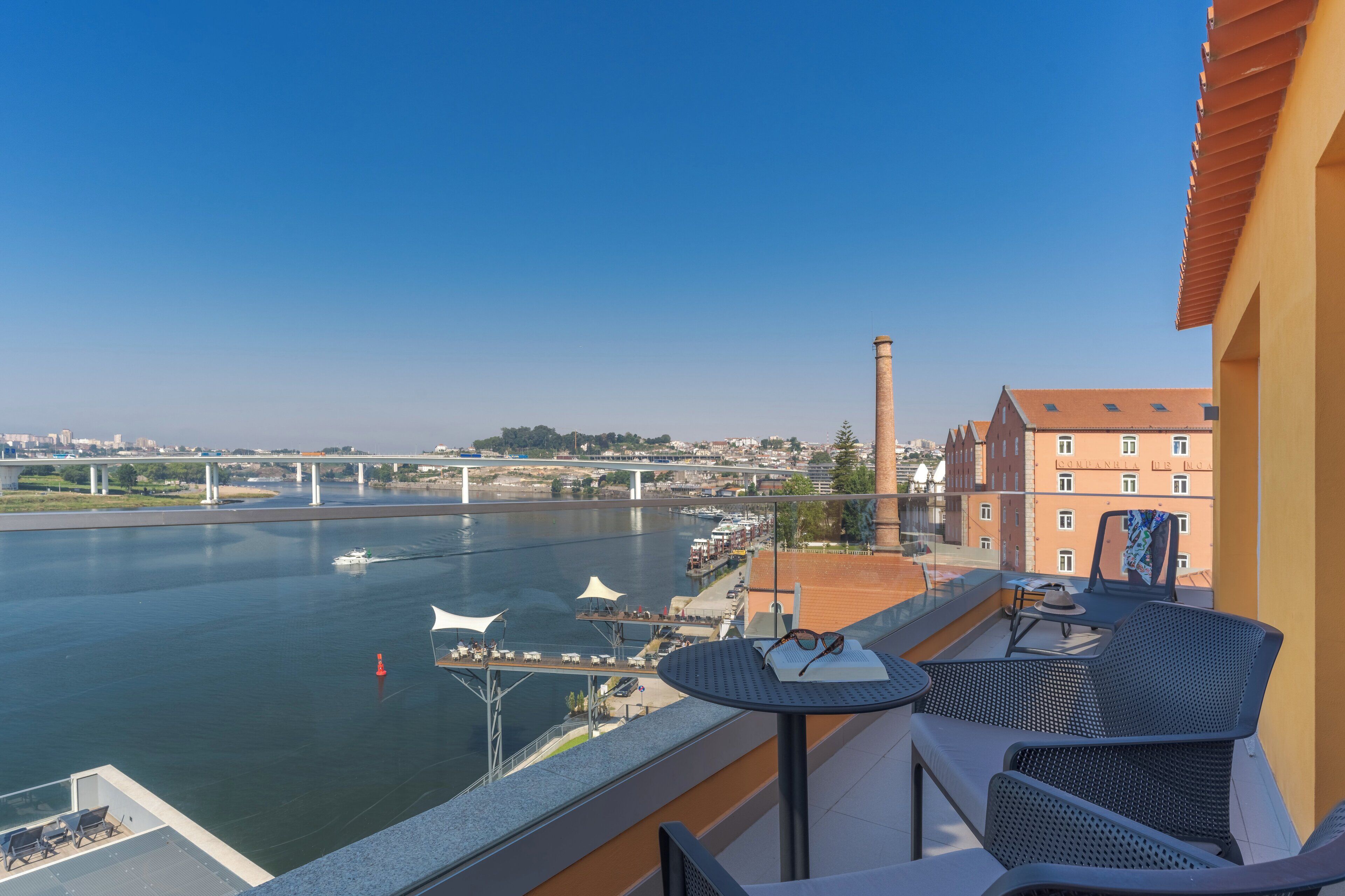 Varanda com mesa e cadeiras, vista para um rio, uma ponte e edifícios ao longe.