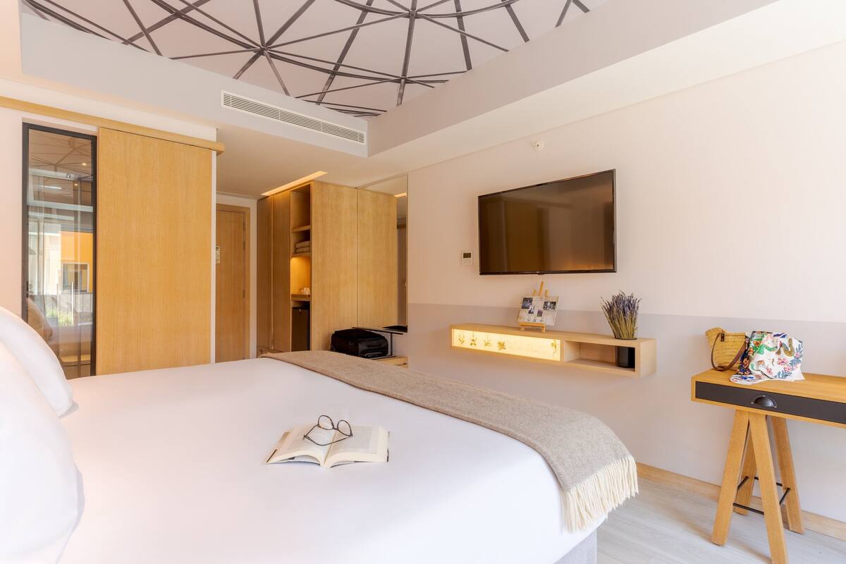 A modern hotel room with a large bed, a TV mounted on the wall, a desk with a lamp, and a wardrobe.
