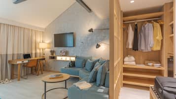 Una habitación de hotel moderna con sofá, escritorio, televisor y vestidor con ropa colgada.