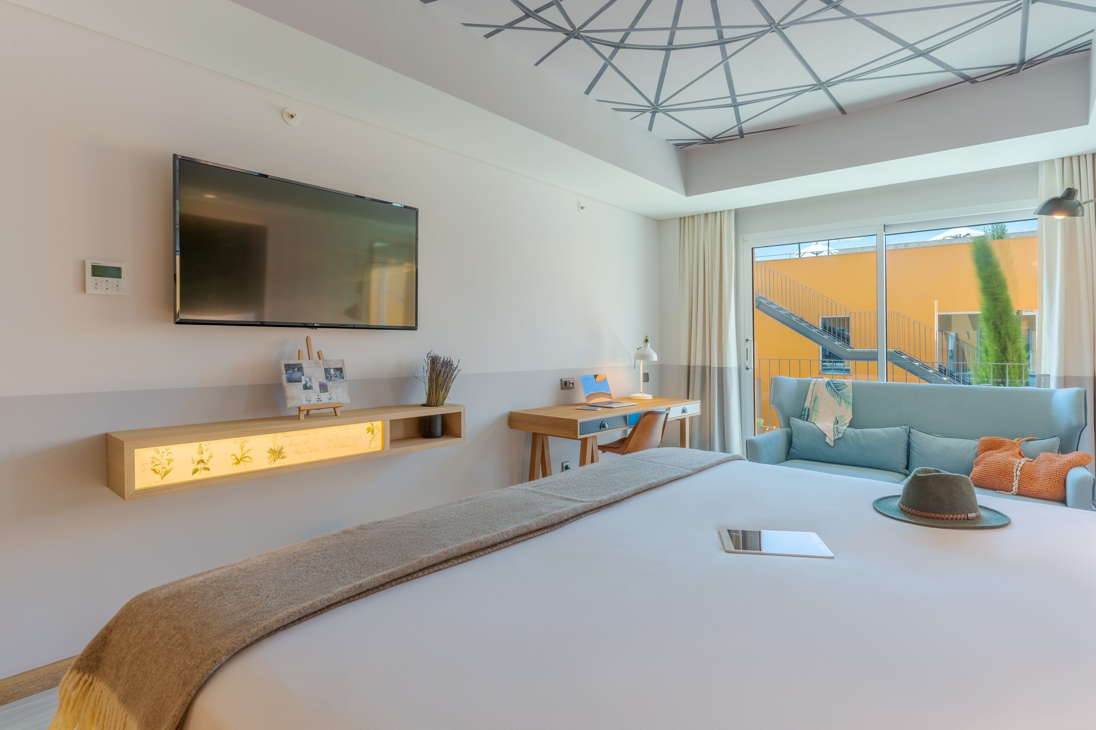 A modern hotel room with a large bed, a desk, a flat-screen TV mounted on the wall, and a view of a building outside.