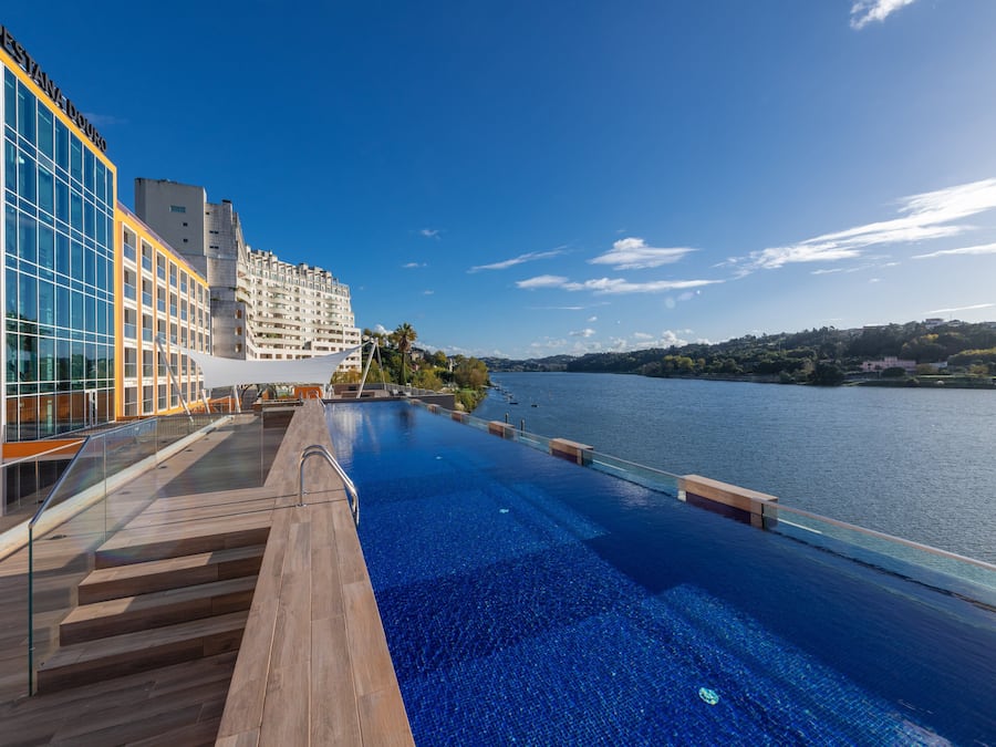 A modern hotel with a large infinity pool overlooking a body of water, a glass facade, and a clear sky.