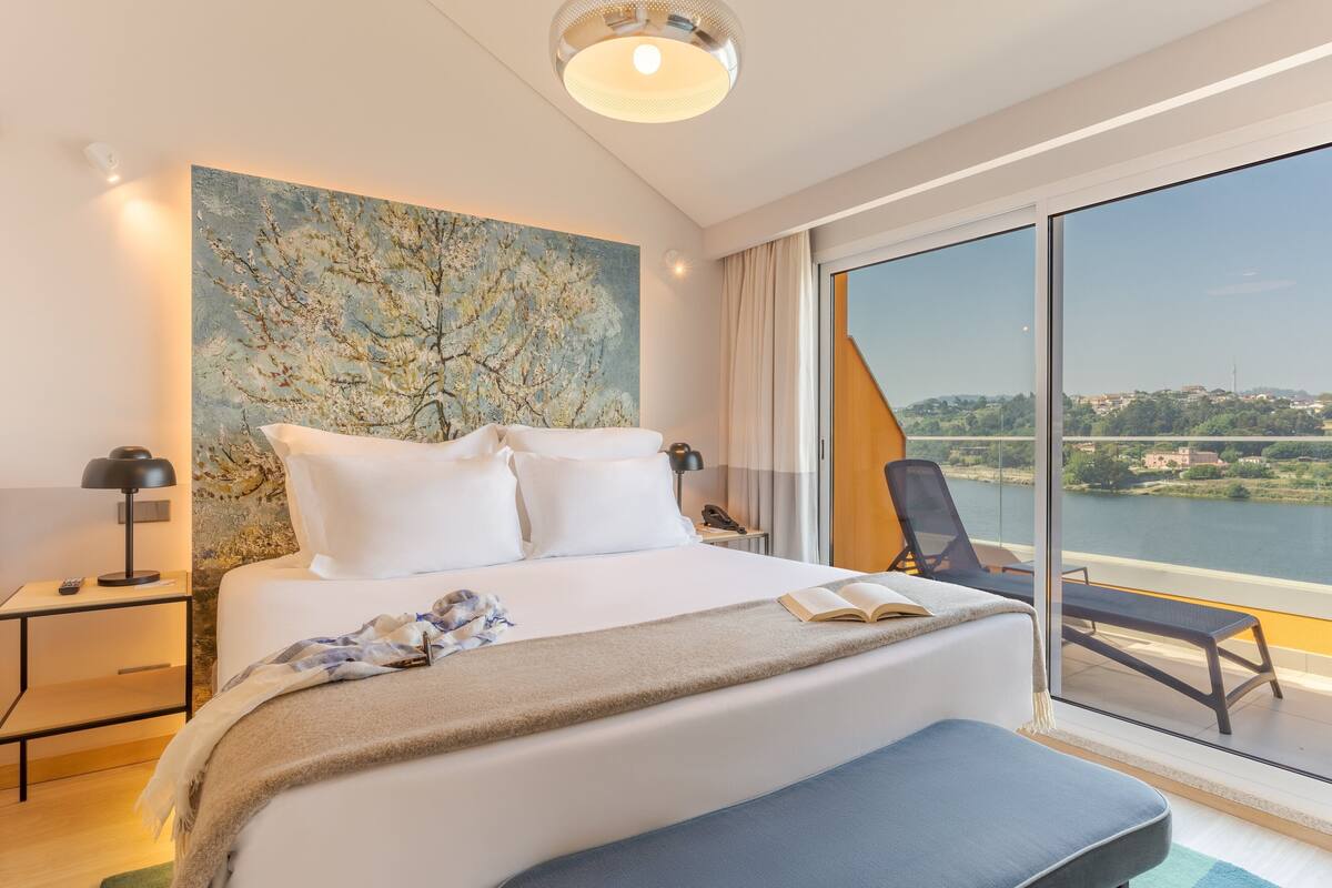 A modern hotel room with a large bed, a painting on the wall, a balcony with a view of a river and cityscape.