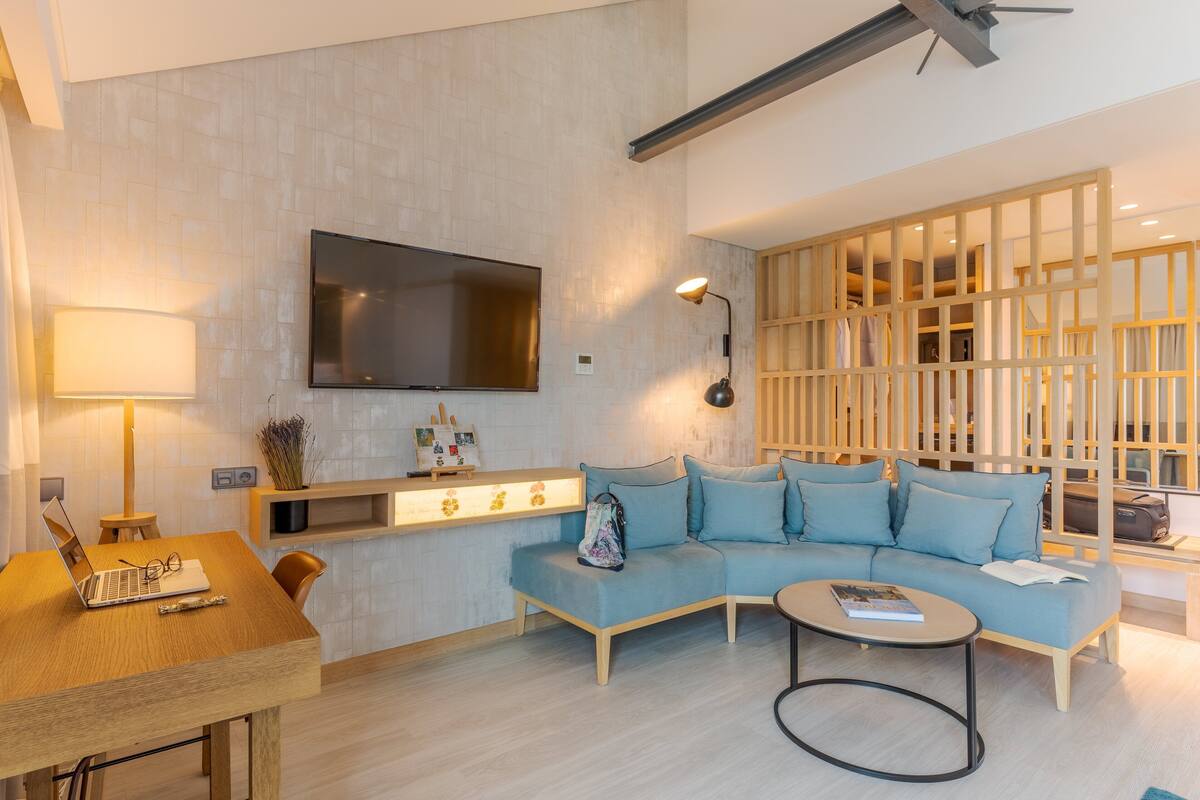 A modern living room with a wooden sofa, a TV mounted on the wall, a desk with a laptop, and a lamp.