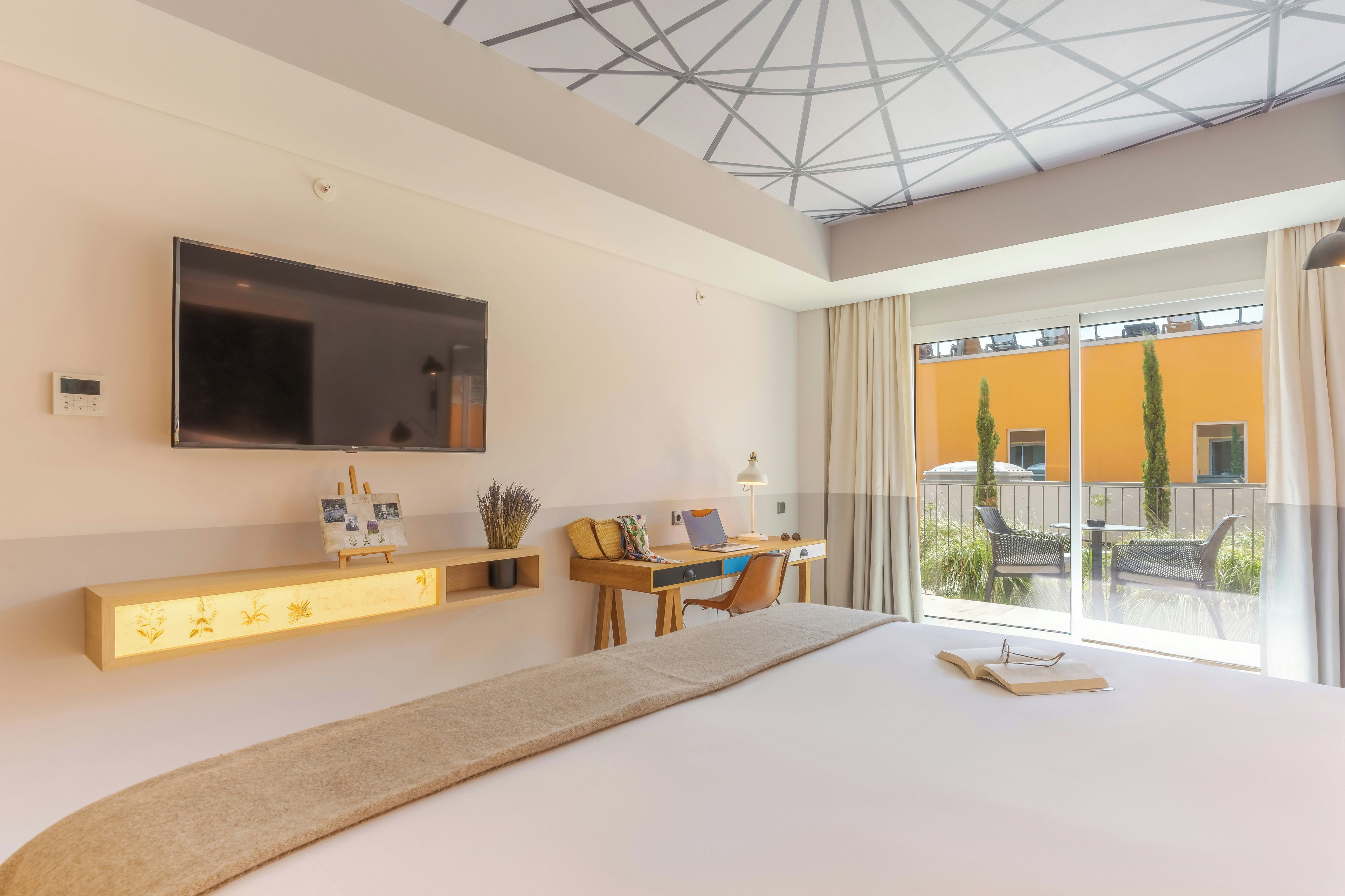 Ein modernes Hotelzimmer mit einem großen Bett, einem Schreibtisch mit Laptop, einem flachbildfernseher an der Wand und einem Balkon mit Gartenmöbeln.