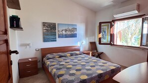 A bedroom with a wooden bed, a nightstand, a chair, and a desk. There are two wall-mounted air conditioning units and a window with a view of greenery. - Villa Marina on the waterfront I.U.N. code R9661 (Magomadas)