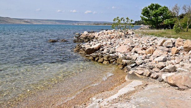 A rocky shoreline with clear water and a distant view of a hilly landscape.