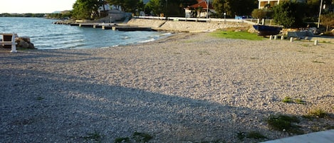 A pebbled beach with a stone wall, a pier, and buildings in the background.