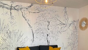 Un salón moderno con un sofá oscuro, cojines amarillos, una mesa de centro redonda y una pared con un mural botánico.