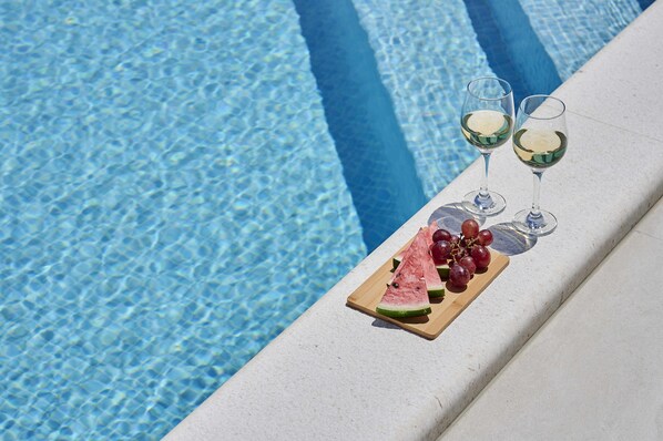 A poolside setting with a wooden board holding watermelon slices and grapes, and two glasses of white wine.