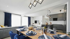 A modern dining area with a long table set for four, a kitchen in the background, and a living area with a sofa and curtains. - Blue fantasy is new modern villa 5 min from the beach! (Kaštel Novi)