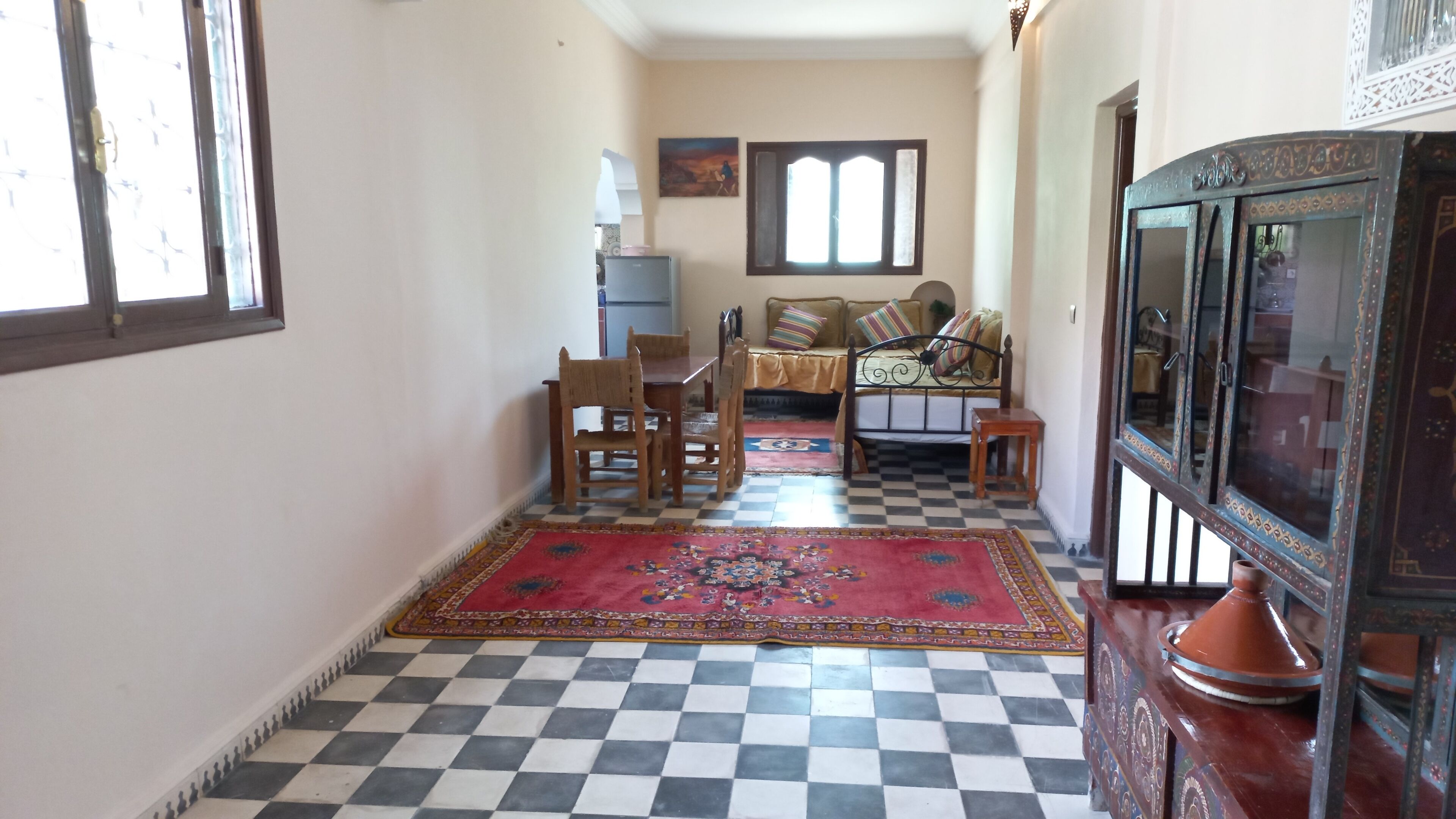 Un couloir doté d’un sol à carreaux, une table en bois avec des chaises, un canapé avec des coussins, un tapis à motifs et un meuble décoratif.