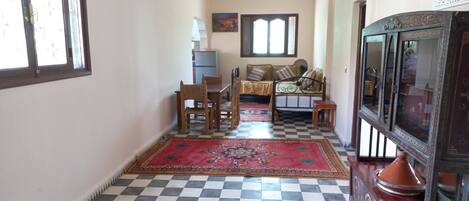 Un couloir doté d’un sol à carreaux, une table en bois avec des chaises, un canapé avec des coussins, un tapis à motifs et un meuble décoratif.