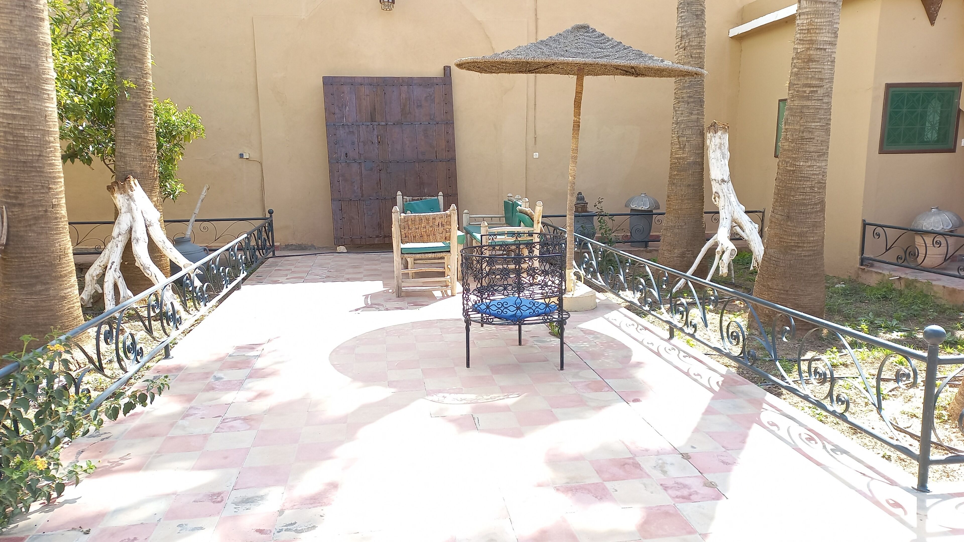 A courtyard with a tiled floor, a wooden door, a metal chair, and a woven umbrella.