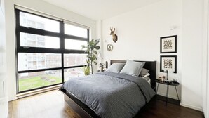 A bedroom with a large window, a bed, a plant, and wall decorations. - London Warehouse One Bedroom Apt (London)