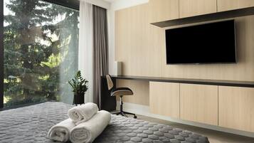 A modern hotel room with a large bed, a flat-screen TV mounted on the wall, a desk with a chair, and a view of trees through the window.