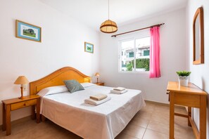 A bedroom with a wooden bed, two bedside tables, a nightstand, a window with a pink curtain, and a small table with a plant.
