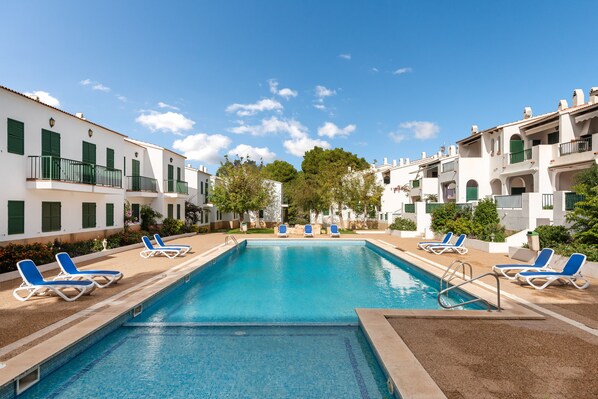 A rectangular swimming pool with blue water, surrounded by white buildings with green shutters and balconies, and blue lounge chairs along the poolside.