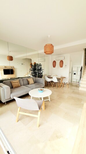A modern living room with a grey sofa, a round coffee table, and a dining area with white chairs and a small table. - Ocean Calma Apartment 5 / Fast WIFI / huge pool and kids pool  (MOGAN)