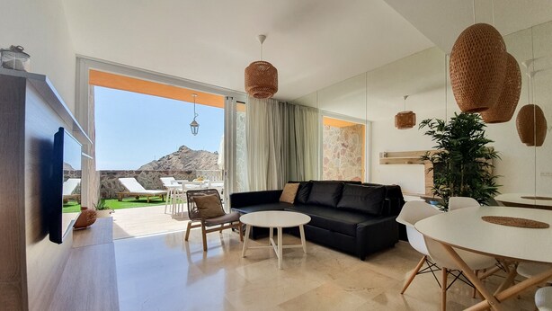 Una sala de estar moderna con un sofá negro, una mesa de centro blanca y una mesa de comedor. Hay un televisor montado en la pared, un amplio ventanal con vistas al exterior y una pared de piedra.