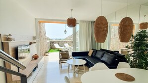 Una sala de estar moderna con un sofá negro, sillas blancas y una mesa de comedor redonda. Se observa una escalera a la izquierda y vistas al exterior a través de un ventanal.