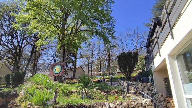 A landscaped garden with a large tree, stone wall, and a building with a white facade. - Kamea Club La Gironie (Turenne)