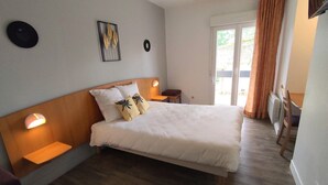 A bedroom with a wooden headboard, a bed with white bedding, two bedside tables with lamps, a small stool, and a view of the outdoors through a sliding door. - Kamea Club La Gironie (Turenne)
