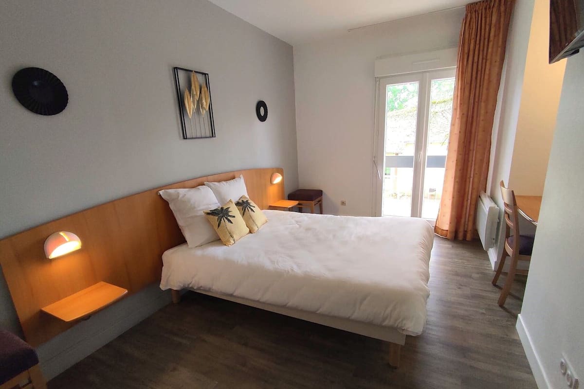 Een slaapkamer met een houten hoofdbord, een bed met wit beddengoed, twee nachtkastjes met lampen, een klein krukje en uitzicht op het buitenlands landschap door een schuifdeur.