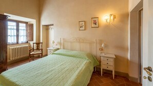 Un dormitori amb un llit, dues taules de nit, una cadira, un radiador, una finestra amb cortines i làmpades penjades a la paret.