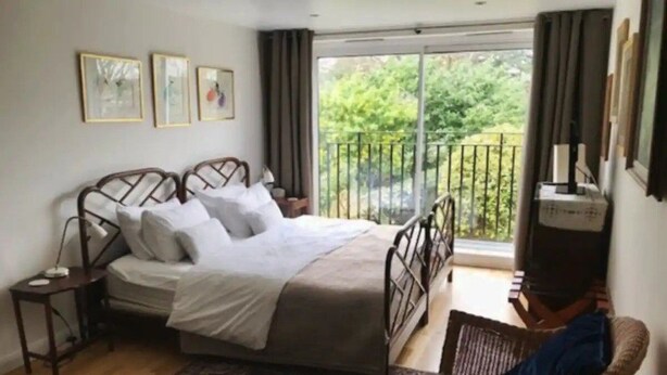 A bedroom with a bed, a chair, a small table, and a balcony with a view of greenery.