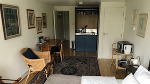A hotel room with a bed, a dining table with chairs, a kitchen area, and a small desk with a lamp.