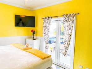 A bright yellow room with a bed, a small white bedside table with a vase of flowers, and a TV mounted on the wall. - Ocean Private Retreat 3 Apt Suites w Pool Sleeps 18 (Nassau)