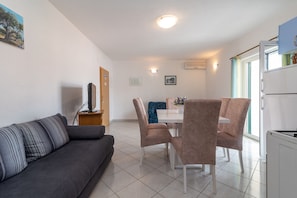 Un salon moderne avec un canapé gris, un coin repas avec une table et des chaises blanches, et un espace cuisine équipé d’un réfrigérateur.