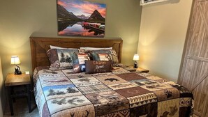 A bedroom with a wooden headboard, a quilted bedspread with wildlife prints, two bedside lamps, and a painting of a mountain landscape above the bed.