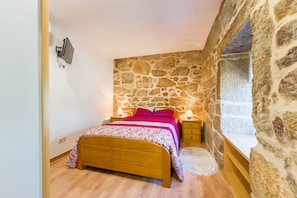 A bedroom with a stone wall, a wooden bed, a nightstand, and a window with a view.