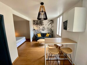 Un espacio habitable compacto con sofá, una mesa de comedor con sillas, una lámpara colgante y una pared decorada con un mural tropical.