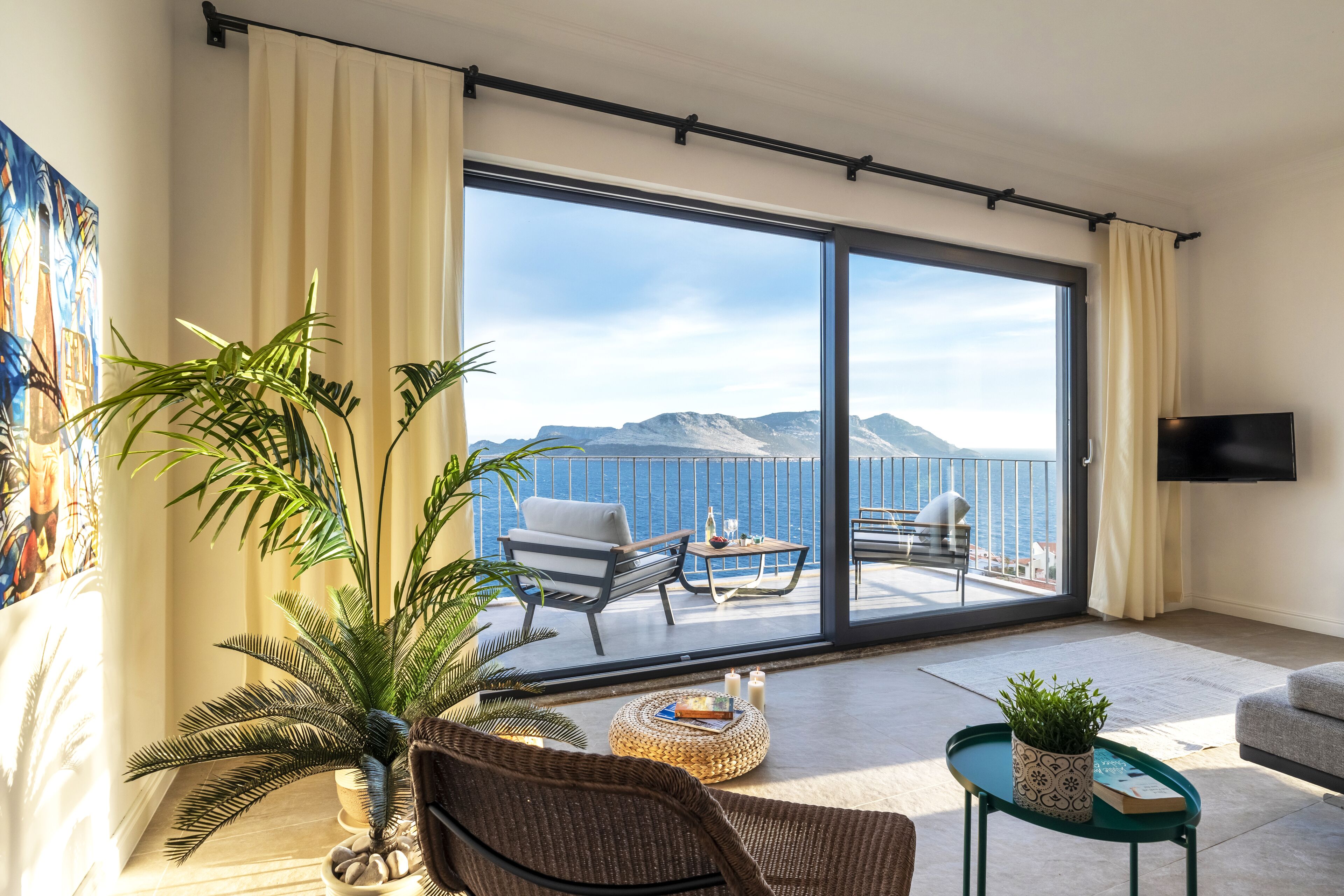 Un salon doté d’une grande porte-fenêtre coulissante donnant sur un balcon avec vue sur la mer et les montagnes. On y trouve deux fauteuils, une table basse et un téléviseur fixé au mur.
