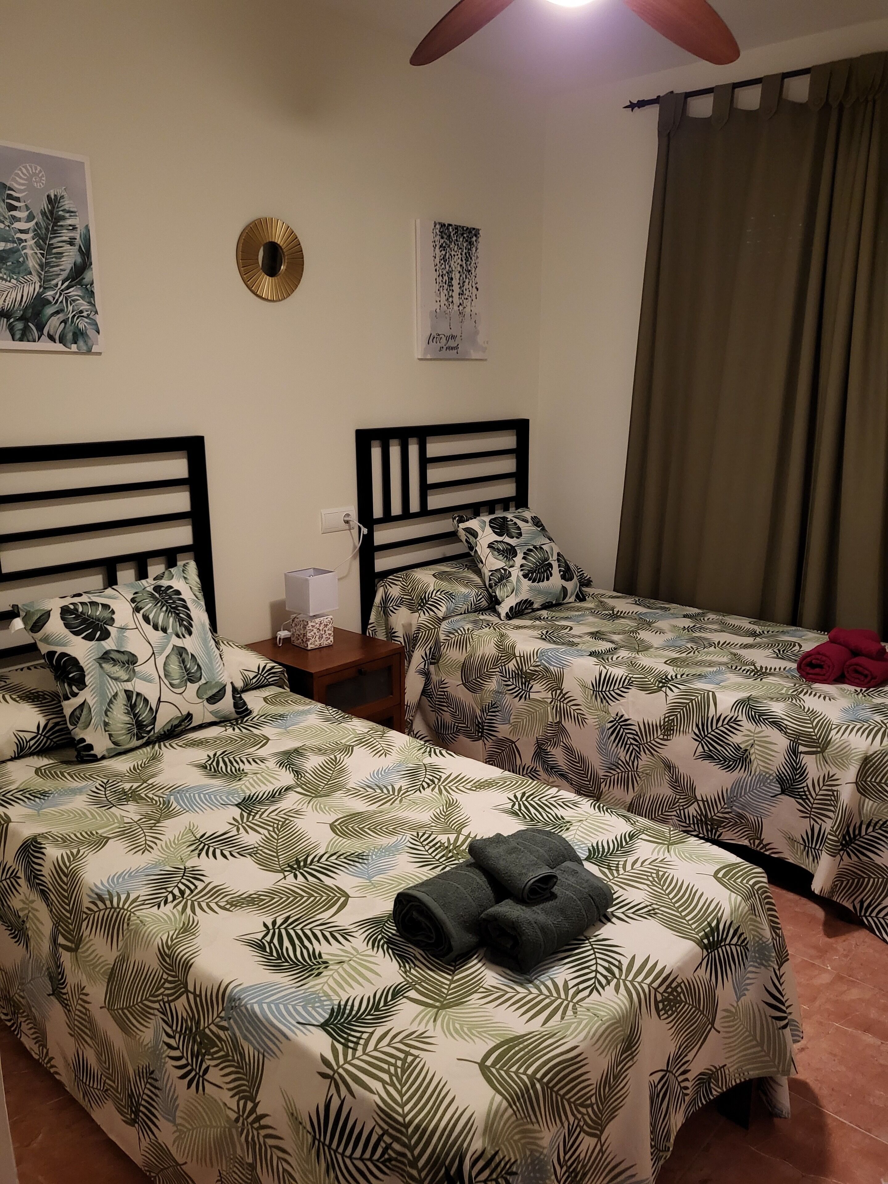 Quarto com duas camas, cada uma com colcha estampada em folhagem, uma mesinha de cabeceira com luminária, e uma parede com pintura de tema tropical e uma citação emoldurada.