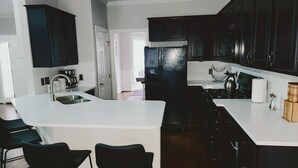 A modern kitchen with dark cabinets, a white countertop, and a stainless steel sink. There are two black bar stools and a refrigerator with a water dispenser. - Remodeled ranch style home with Firepit conveniently located (Raleigh)