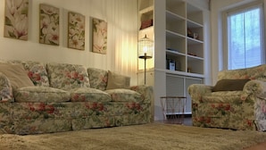 En stue med blomstermønstrede sofaer, en lurvet måtte og et reolsystem med diverse genstande.