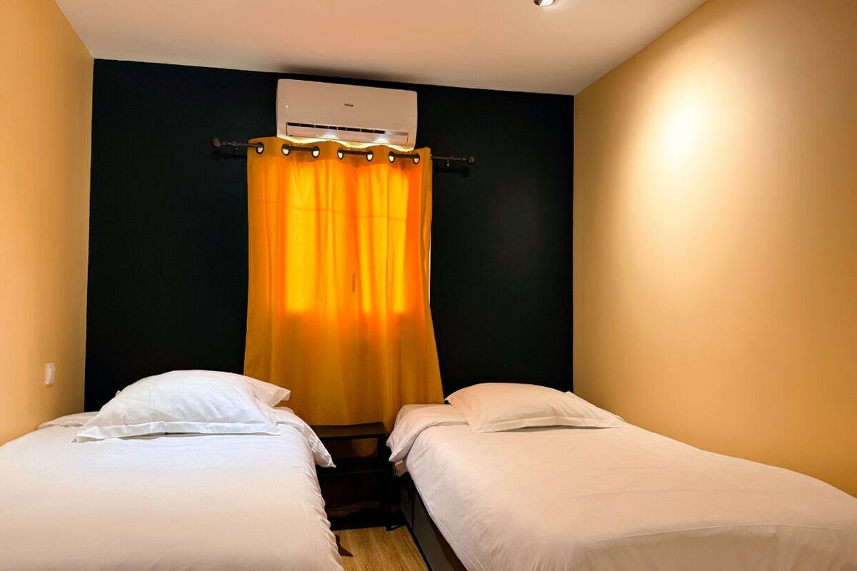 A small, single-room hotel room with two single beds, a dark-colored wall, and a ceiling-mounted air conditioning unit.