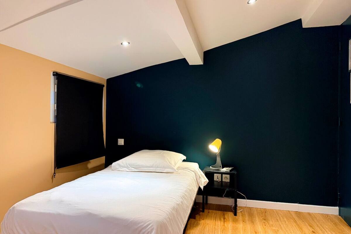A bedroom with a bed, a bedside table with a lamp, and a window with blinds.