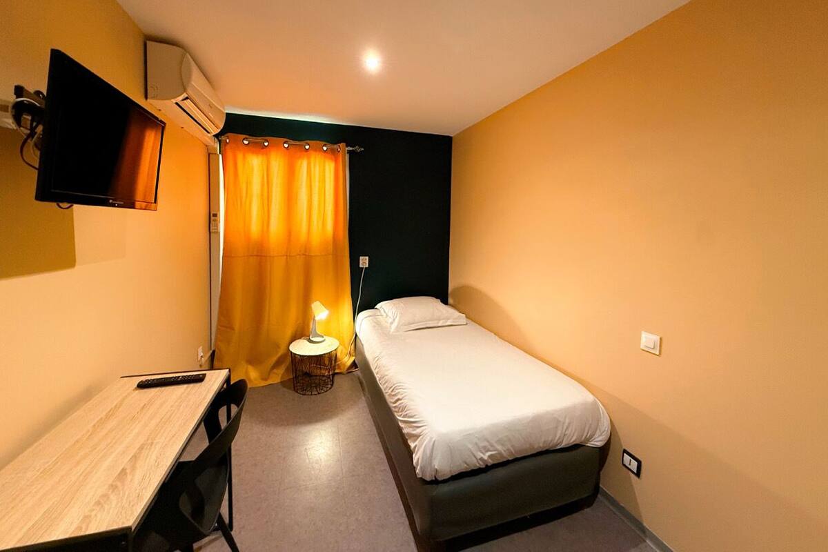 A small, single-room hotel room with a bed, a desk, a chair, a TV, and a window with curtains.